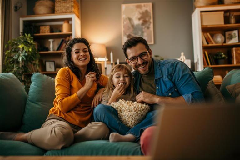 A family of three people sitting on a sofa at home with a lamp in the background. The young girl sitting in the middle eating popcorn, watching TV