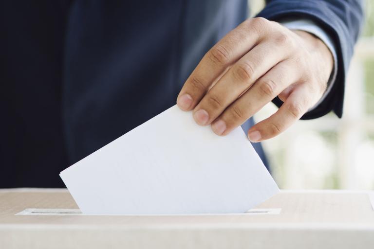 A close up of a male hand putting a piece of paper into the opening of a ballot box.