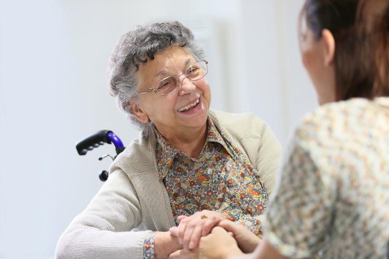 A photograph of an elderly lady in a wheelchair smiling at a carer who is holding her hand. The carer has her back to the camera.