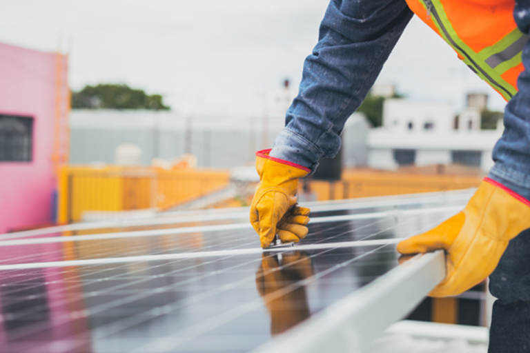 A worker in an orange high visibility vest and yellow gloves completing the installation of a solar panel. 