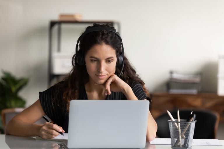 A woman wearing over-ear headphones looking at a laptop. She is holding a pen and has a piece of paper in front of her.