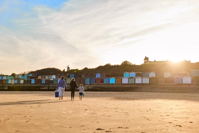 A parent and 2 children walking across sand towards some beach huts 