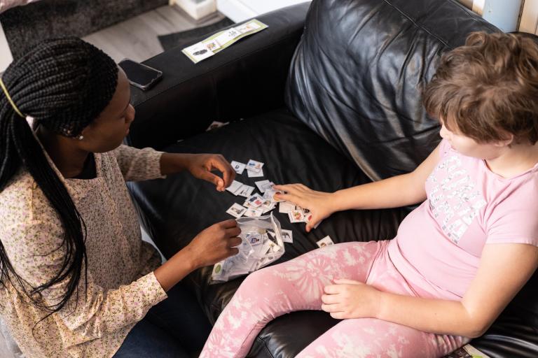 A woman is kneeling before a black sofa with a young child wearing pink pajamas sitting on the sofa and they are sorting through some bits of paper together