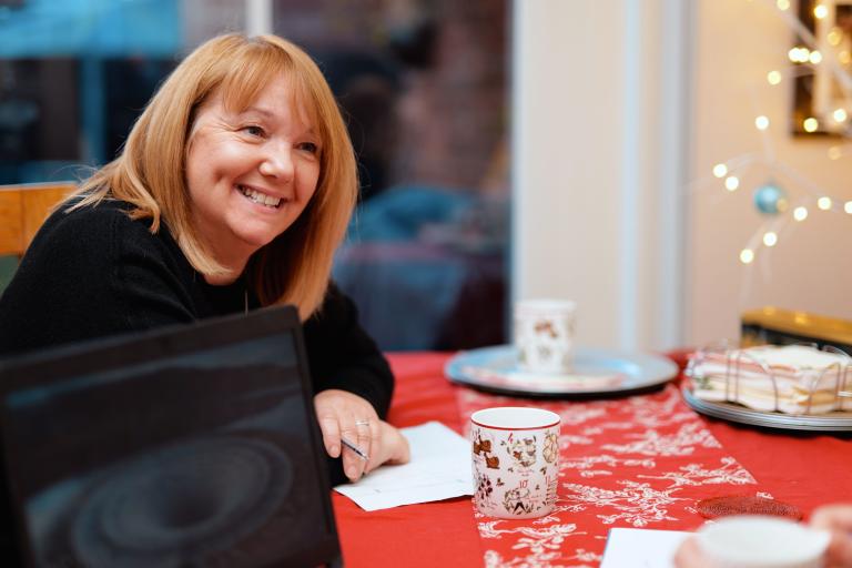 A woman with auburn hair is smiling, sat at a table with red table cloth and winter decorations in the background 