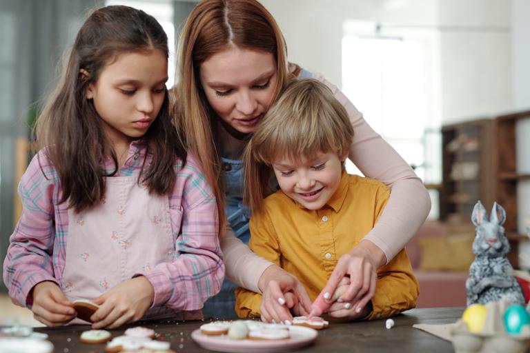 A small boy with blond hair and a older child with long brown hair are making cookies, a woman has her arms wrapped around the boy helping him add decoration 