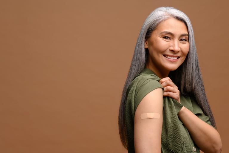 Older woman smiles to camera while exposing plaster over arm after vaccination.