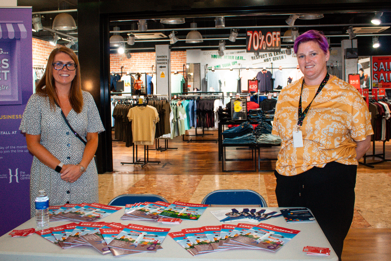 Two women standing behind a desk with leaflets on at a careers fair event.