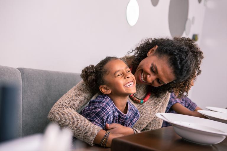 Young child smiling at the dinner table with her foster carer wrapping her arms round her.