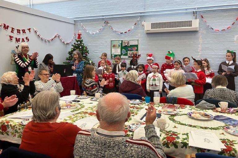 A group of young people singing whilst a crown of older residents watch on