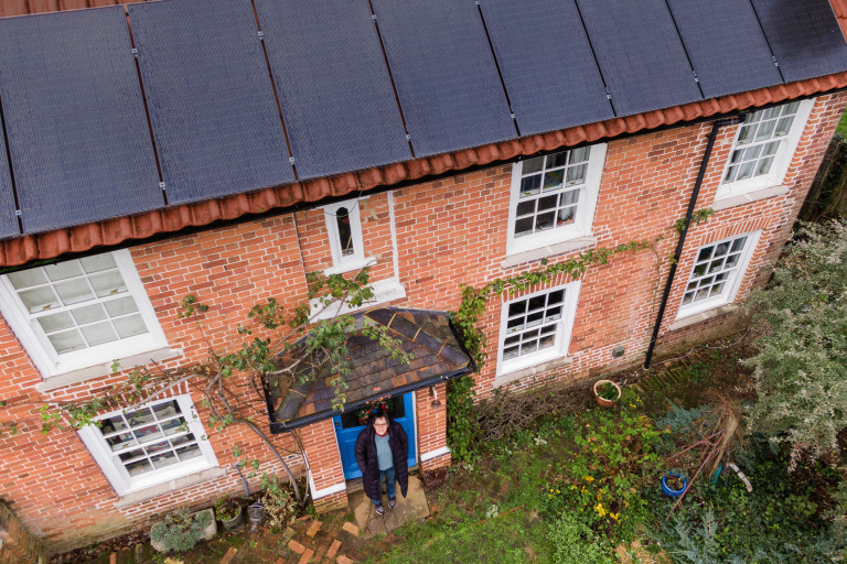 Woman stood outside her front door stares up to an aerial view of her house.