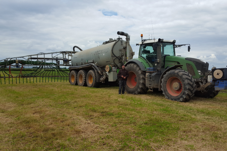 A farmer stands in front of a tractor and plough in the middle of a field.