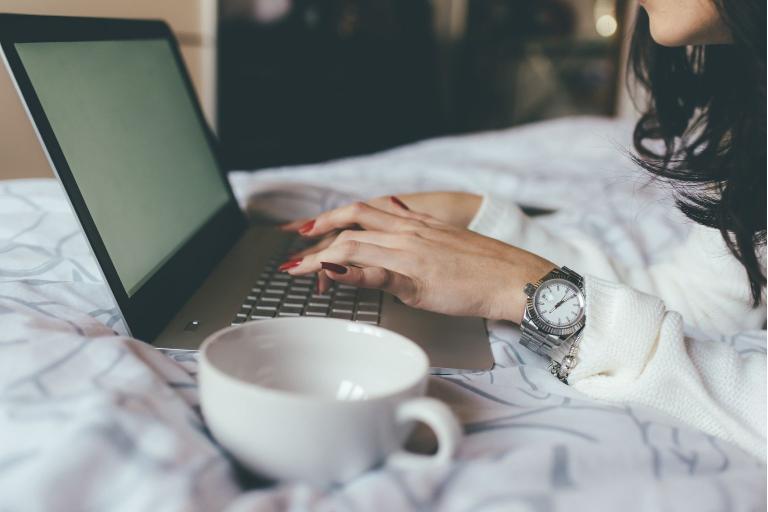 A woman is shown laying on a bed while typing on a laptop. A mug is on the side next to the laptop.