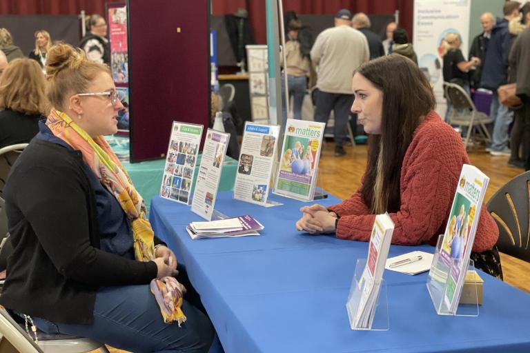 Two women sit across a table in an exhibition hall and speak.