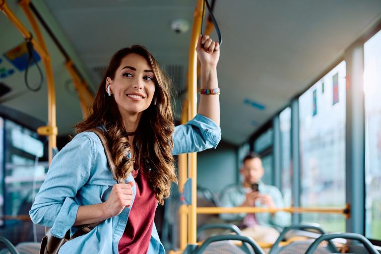 A smiling young woman standing on a bus, holding on to a rail.