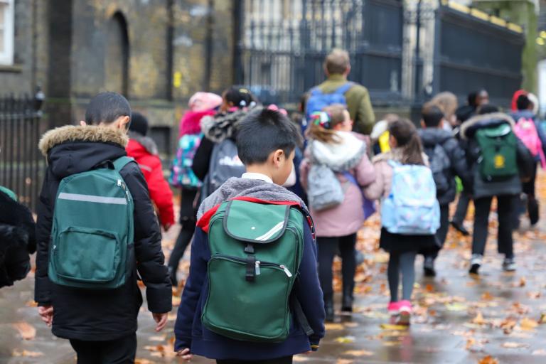 A group of children photographed from behind while walking to school, wearing uniform and backpacks.