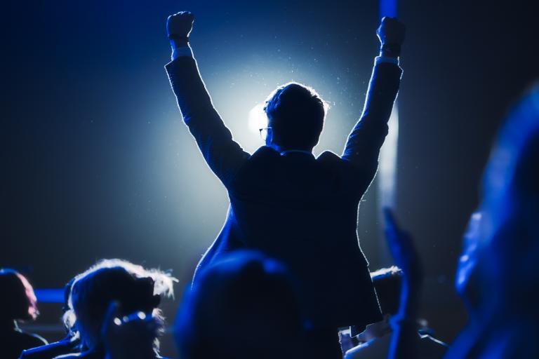 Young man in business attire silhouetted against spotlight as he stands up to cheer.