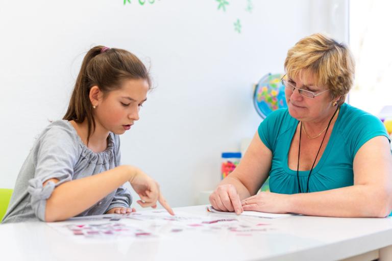 A teenage girl sitting at a table pointing to something on a worksheet while an older lady with a landyard looks down.