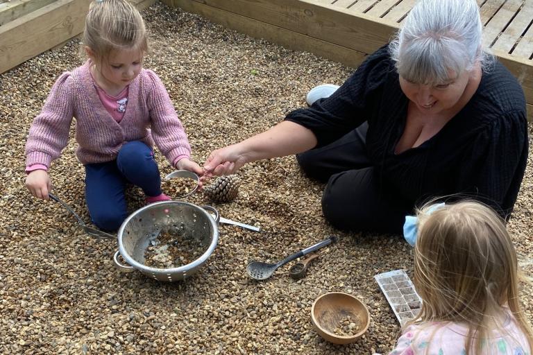 An adult and two children play in a gravel pit and fill up a saucepan.