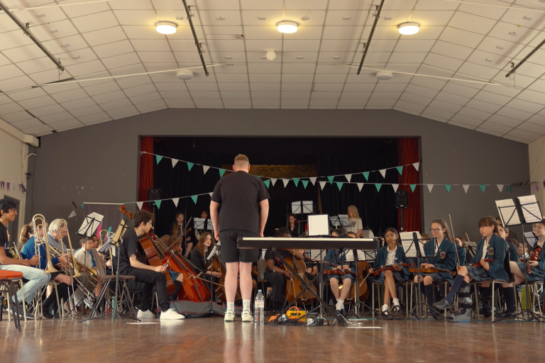 Wide shot of an orchestra of students in an assembly hall with instruments.