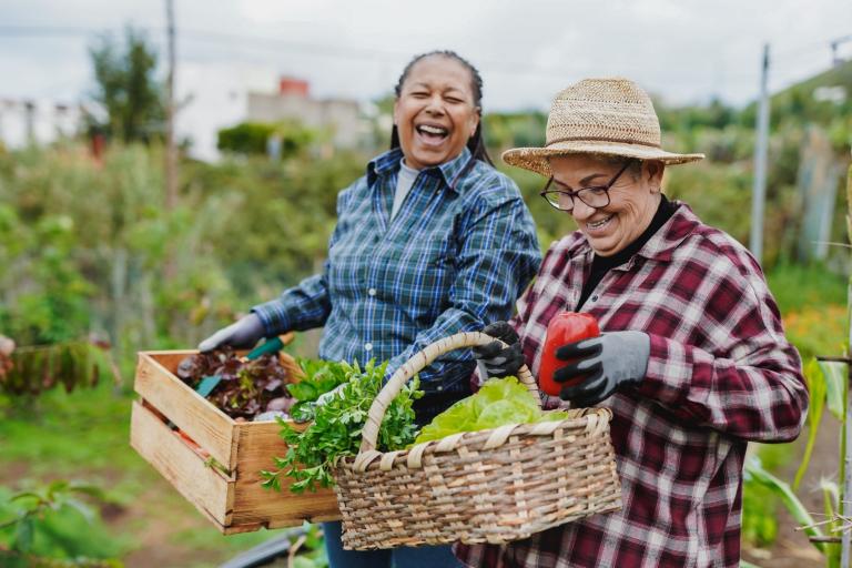 Two women smiling and carrying baskets of garden produce.