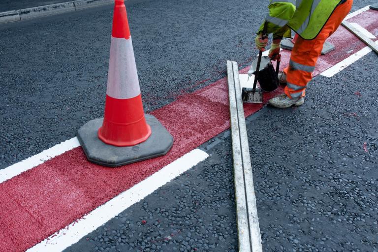 A road worker in high-visibility clothing lays down some red asphalt on a piece of road next to an orange traffic cone.