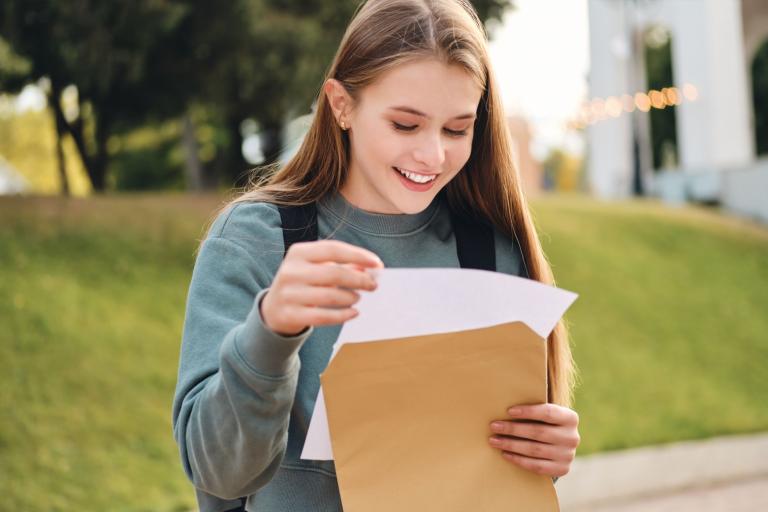 A teenage girl holding an envelope and a piece of paper. She is looking at them and smiling