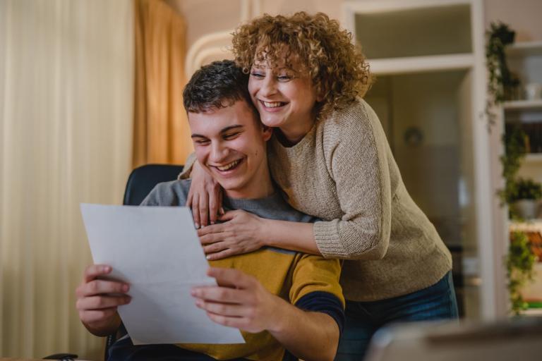 A young man holding a piece of paper is hugged from behind by his mother. Both are smiling.