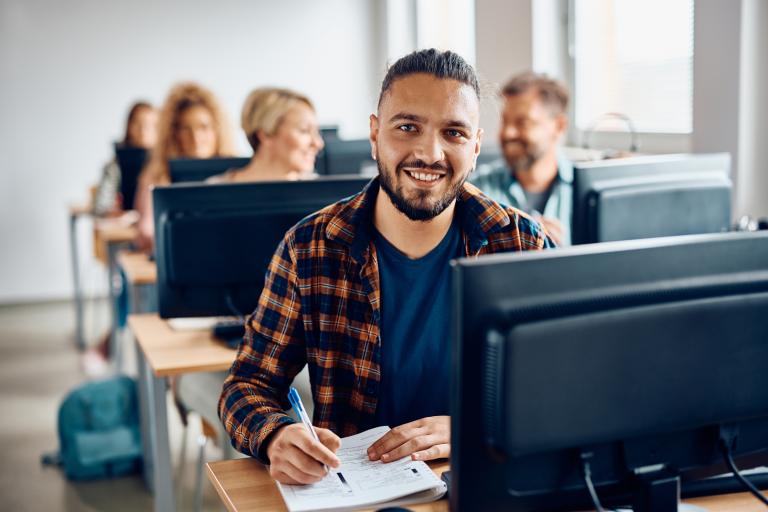 Smiling man sat at t computer and holding a pen against a pad of paper.