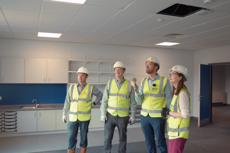 Four people in hi-vis and helmets look to the window in a classroom being built.