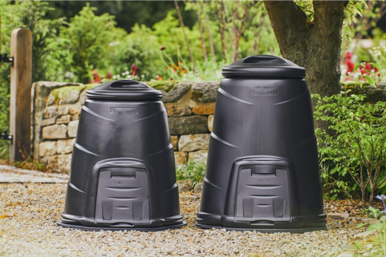 Two black compost bins placed on gravel with a lush garden in bloom in the background.