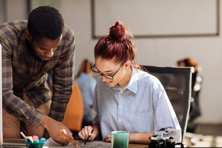 A young female sits at a desk while a male colleague stands next to her looking at papers.