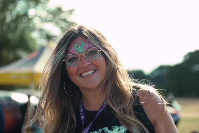 Women with face paint and heart-shaped glasses looks to camera in front of festival tent in a field.