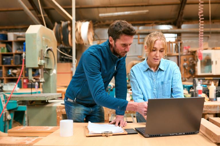 A man and a woman looking towards a laptop screen which is placed on a bench in a carpentry workshop.