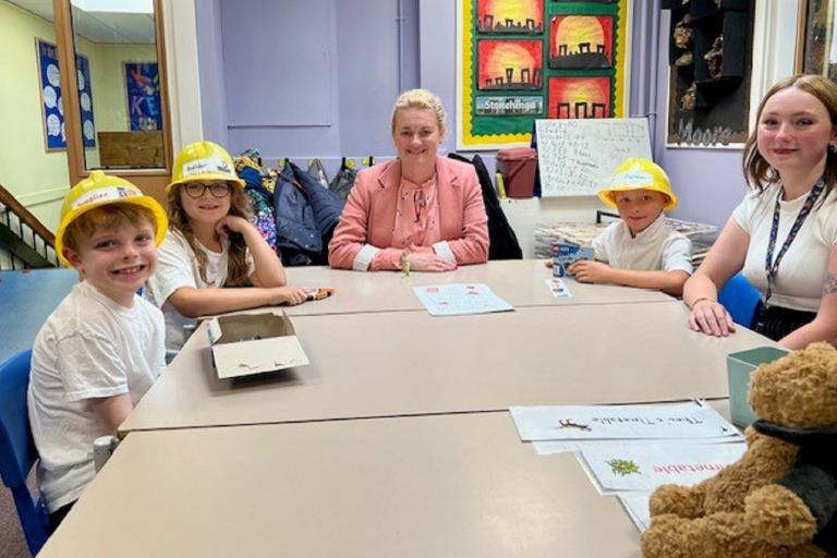 Three children wearing yellow hard hats sitting at a table inside a classroom with two adults.