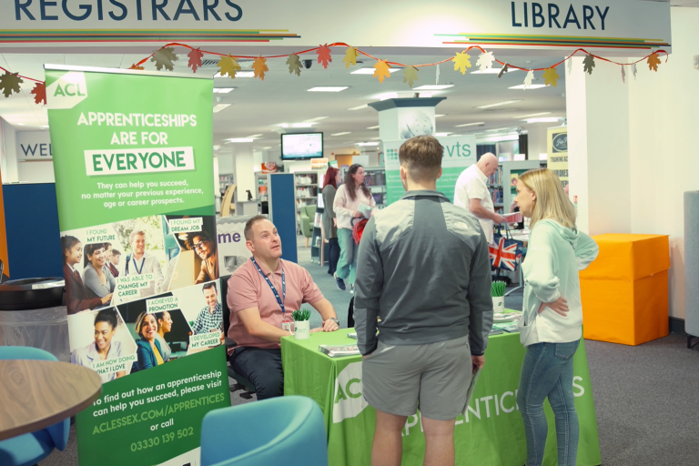 Man sits behind a stall in front of a banner while giving careers advice to a young man.