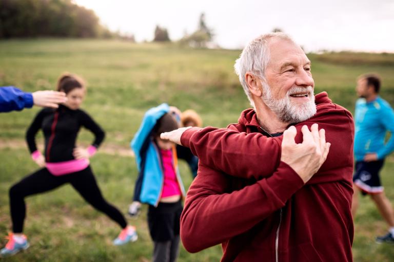 An older gentleman stretching his arms. He is joined by other people dressed in exercise gear preparing to take part in an outdoor activity.