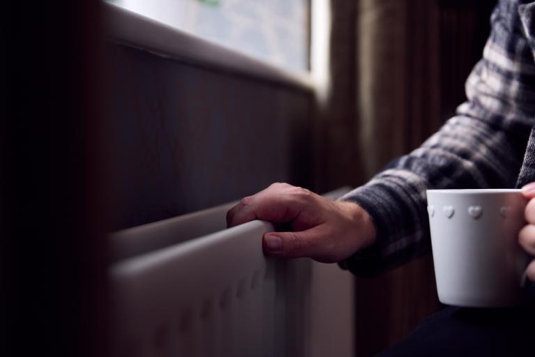 Man in sweater feels radiator with cup of coffee in other hand.