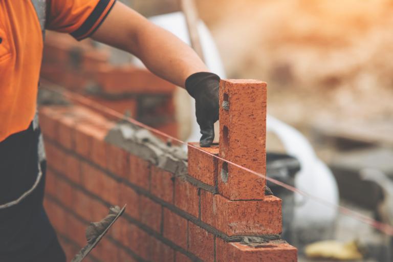 Close-up of worker in hi-vis jacket and gloves laying a brick wall on a sunny day.