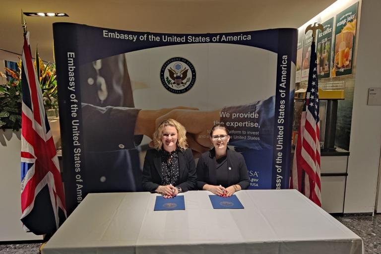 Two women sitting behind a table with official document folders in front of them. Behind is a banner which reads: "Embassy of the United States of America". 