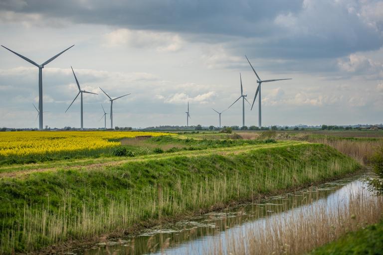 A photograph of a collection of wind turbines located in a field next to a watercourse. 