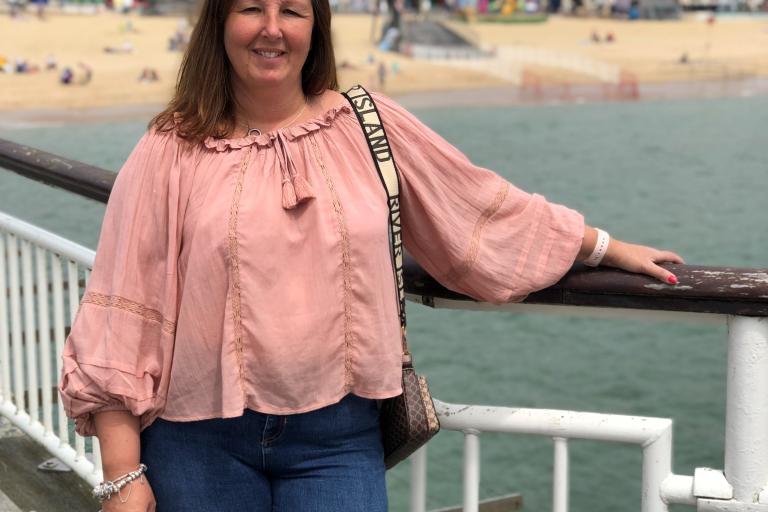 Dionne standing on a pier, with a beach setting behind her. It's a sunny day, the sky is blue. She's smiling, wearing a pink long sleeve blouse and a pair of jeans. 