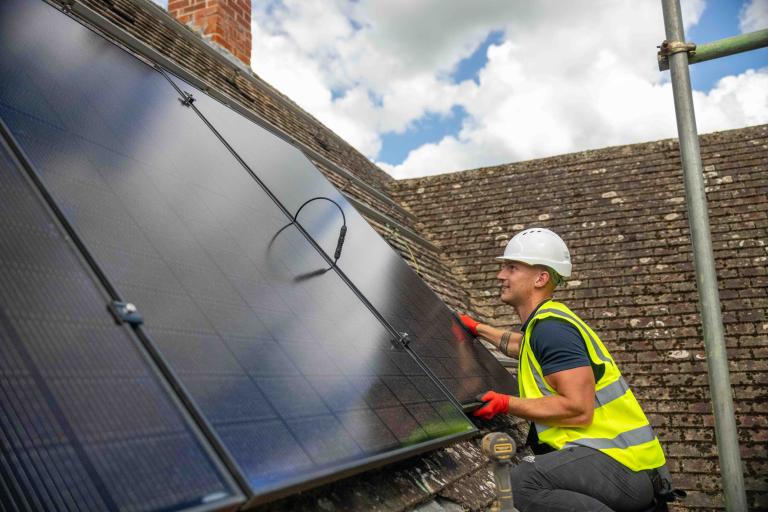 A man wearing a hi-viz vest and hard hat fits a solar panel to a roof.