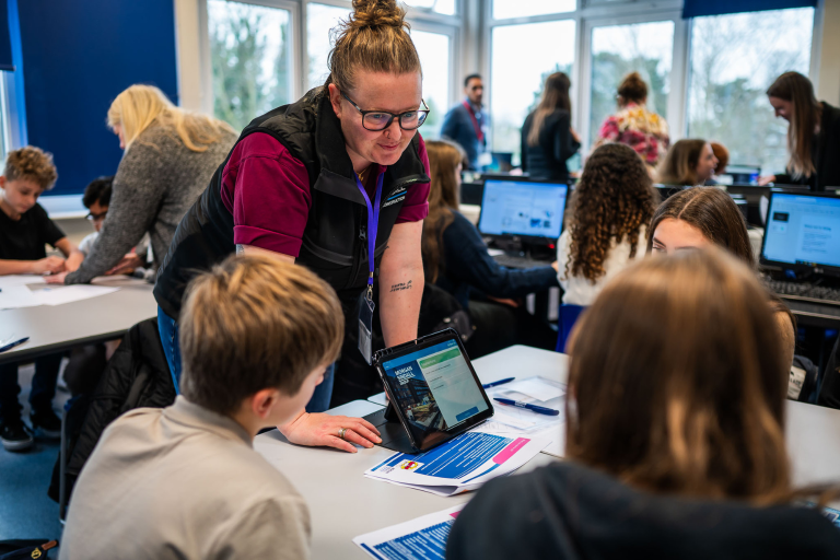 Students gather round tables with tablets and computers to learn about construction.