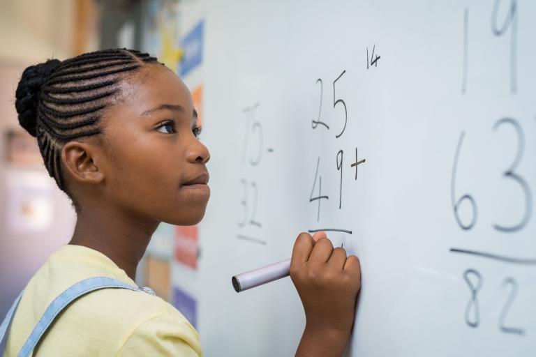 A young female pupil solves an addition sum on a white board with marker pen.