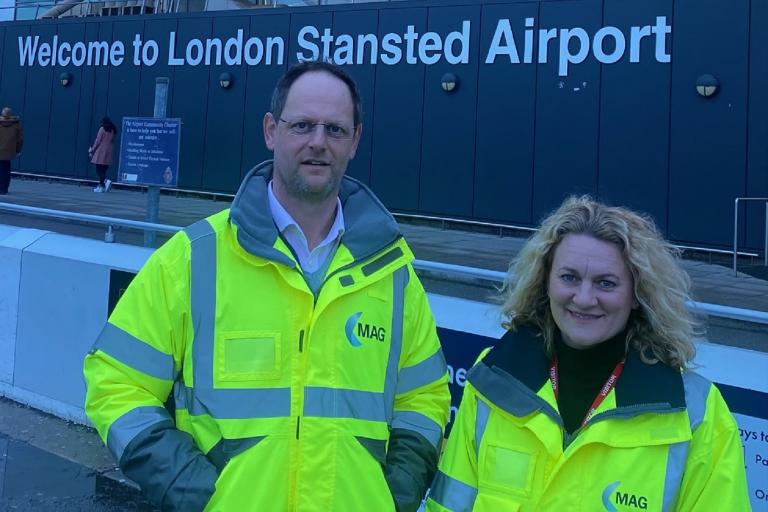 A man and a woman wearing yellow high-visibility jackets standing outside. A sign on a building behind reads: "Welcome to London Stansted Airport".