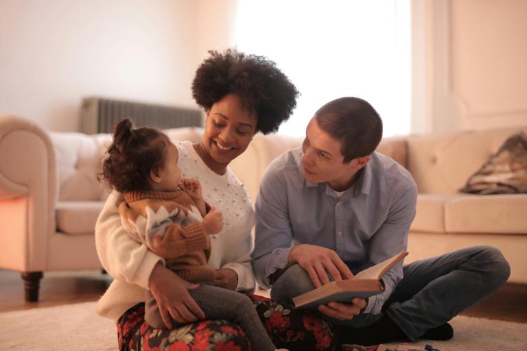 A man and woman sitting on the living room floor with a small girl (aged under 5). The small girl is sitting on the lady's lap, while the man is leaning over to show her the page of a picture book.