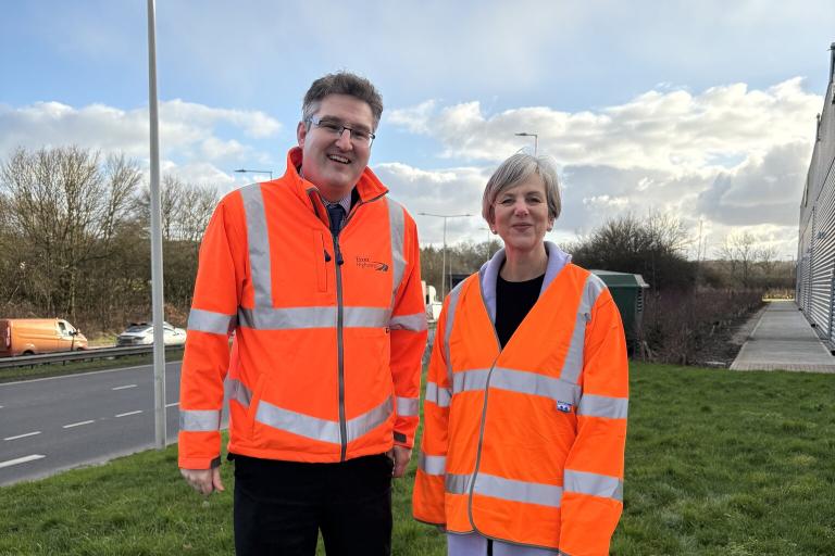 two people standing in high visibility clothing. One is a man and one is a woman. They have roads behind them.